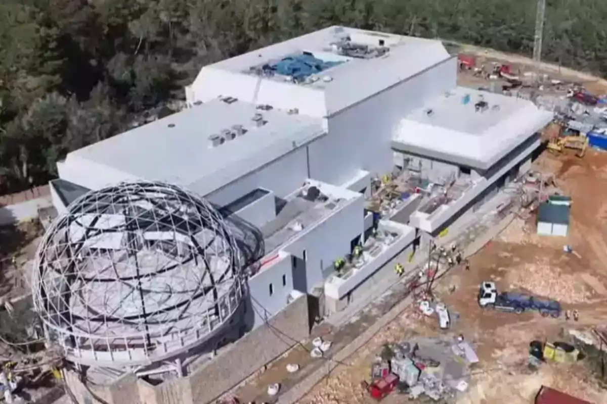 Vista aérea de un edificio en construcción con una estructura esférica metálica en uno de sus extremos rodeado de árboles y maquinaria de obra Vista aérea de un edificio en construcción con una estructura esférica metálica en uno de sus extremos rodeado de árboles y maquinaria de obra