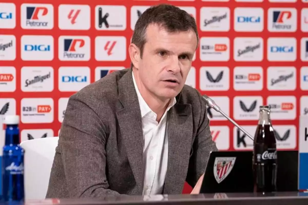 Hombre con saco marrón y camisa blanca hablando en una rueda de prensa con fondo de logotipos y botellas de agua y refresco sobre la mesa Hombre con saco marrón y camisa blanca hablando en una rueda de prensa con fondo de logotipos y botellas de agua y refresco sobre la mesa