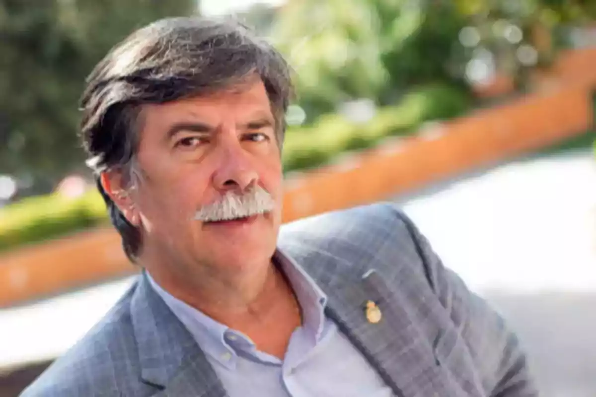 Un hombre con bigote y cabello canoso viste un saco gris y está al aire libre con un fondo desenfocado de vegetación. Un hombre con bigote y cabello canoso viste un saco gris y está al aire libre con un fondo desenfocado de vegetación.