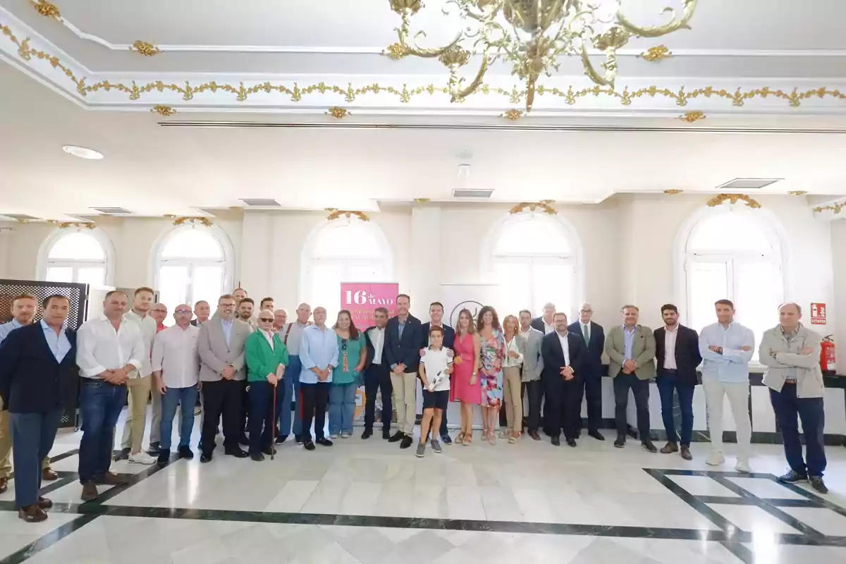 Un grupo de personas posando en un salón elegante con un candelabro dorado en el techo y ventanas grandes al fondo.