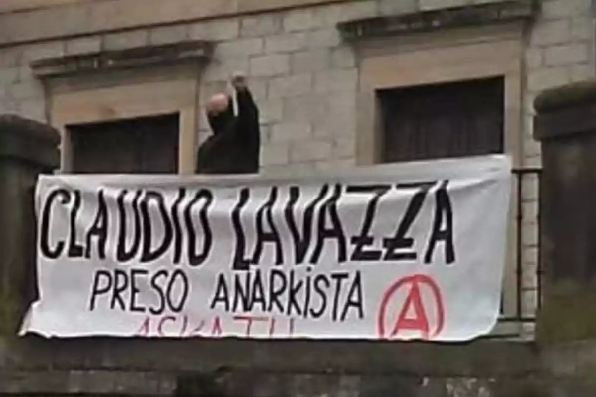 Una persona enmascarada levanta el puño detrás de una pancarta que dice Una persona enmascarada levanta el puño detrás de una pancarta que dice