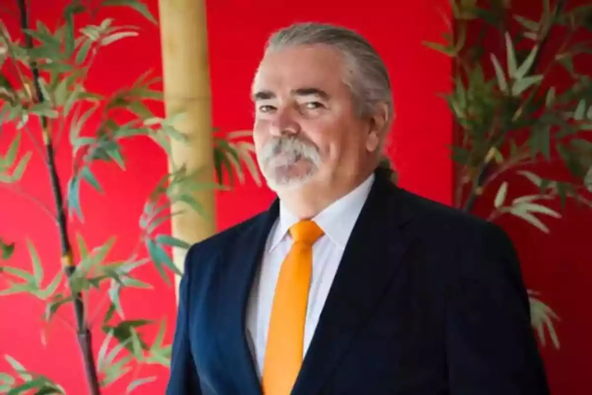 Hombre de cabello canoso y bigote con traje oscuro y corbata naranja posando frente a un fondo rojo con plantas.