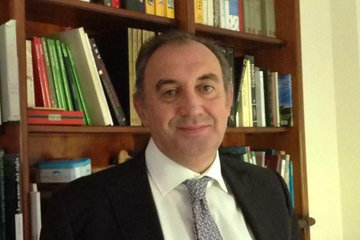 Hombre de traje y corbata sonriendo frente a una estantería llena de libros.