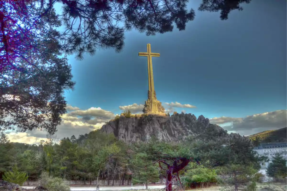 Una gran cruz de piedra se alza sobre una colina rodeada de árboles y cielo azul con nubes. Una gran cruz de piedra se alza sobre una colina rodeada de árboles y cielo azul con nubes.