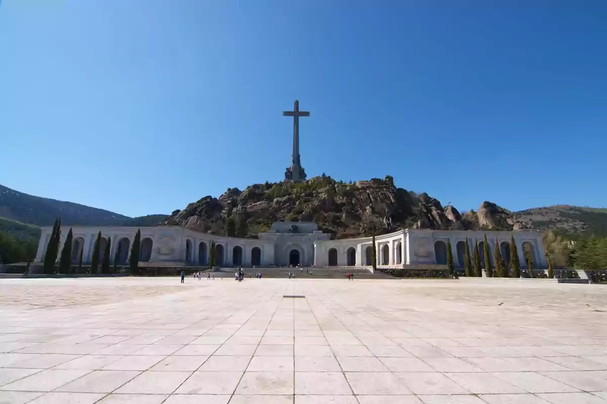 Una gran cruz se alza sobre una colina rocosa con un edificio de arcos en la base y un cielo despejado. Una gran cruz se alza sobre una colina rocosa con un edificio de arcos en la base y un cielo despejado.