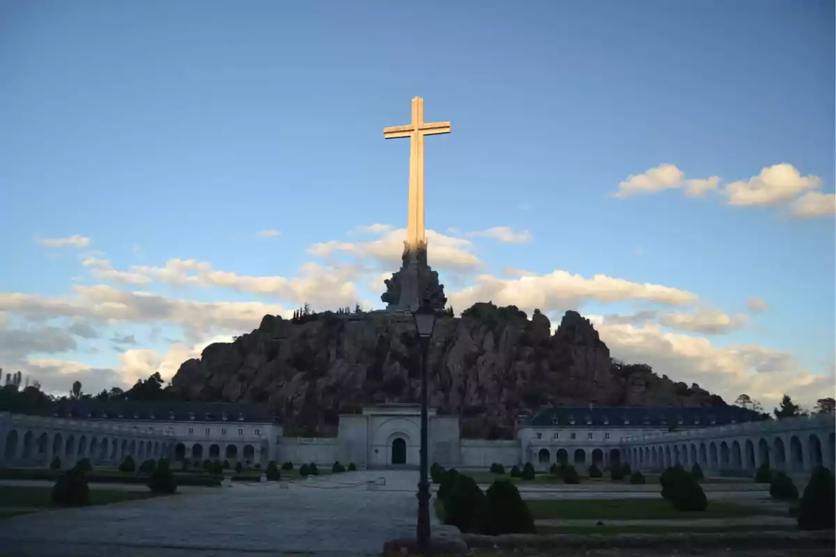 Una gran cruz se alza sobre una colina rocosa rodeada de un edificio con arcos y un cielo azul con nubes.