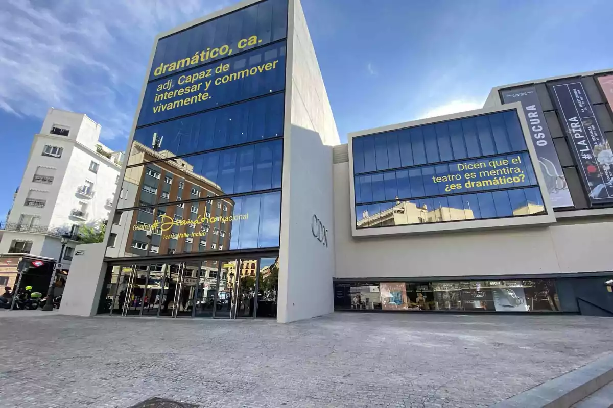 Edificio moderno con grandes ventanales de vidrio que reflejan el cielo y los edificios circundantes, con letreros en amarillo que incluyen definiciones y preguntas sobre el teatro, y la entrada principal del Centro Dramático Nacional.