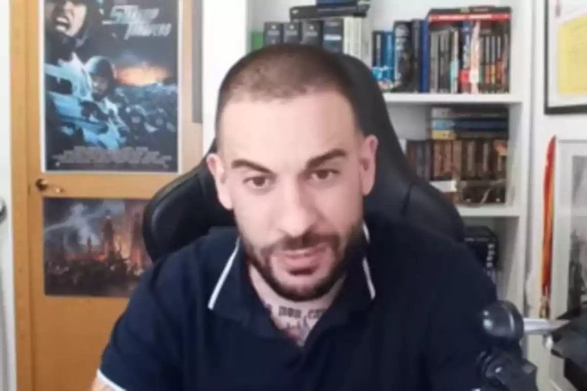 Un hombre con barba y cabello corto está sentado frente a una estantería llena de libros y un póster de película en la pared. Un hombre con barba y cabello corto está sentado frente a una estantería llena de libros y un póster de película en la pared.
