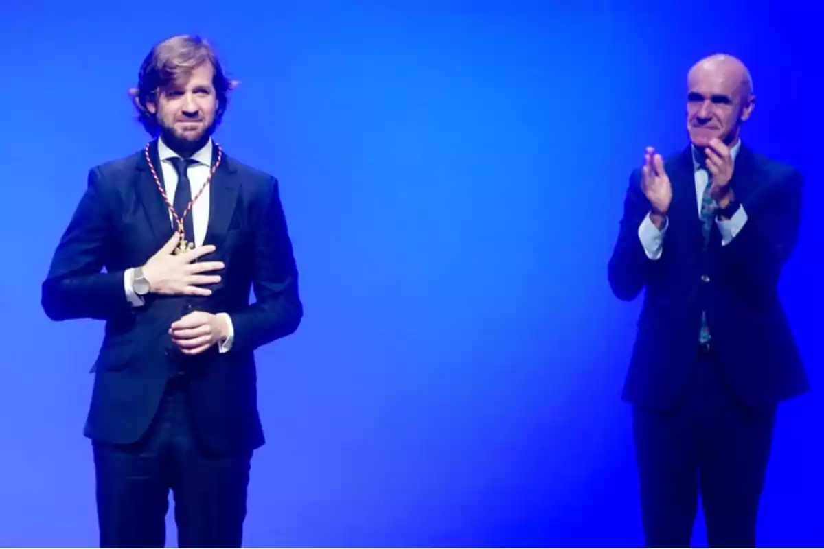 Dos hombres en trajes formales, uno con una medalla y el otro aplaudiendo, en un escenario con fondo azul.