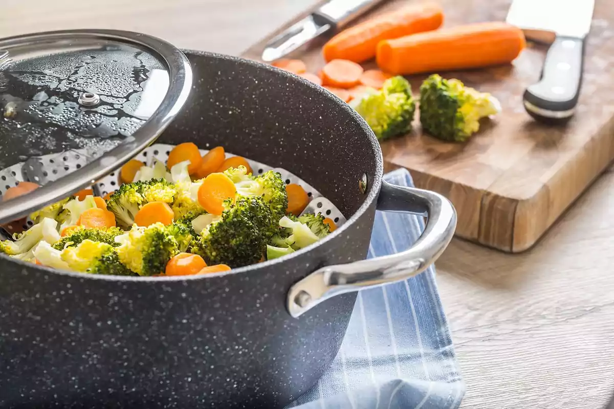 Olla con brócoli y zanahorias al vapor sobre un paño de cocina azul con tabla de cortar y cuchillo al fondo.