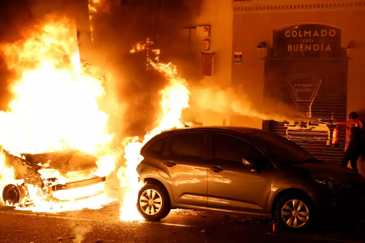 Un coche en llamas junto a otro vehículo mientras una persona intenta apagar el fuego con un extintor frente a una tienda llamada 