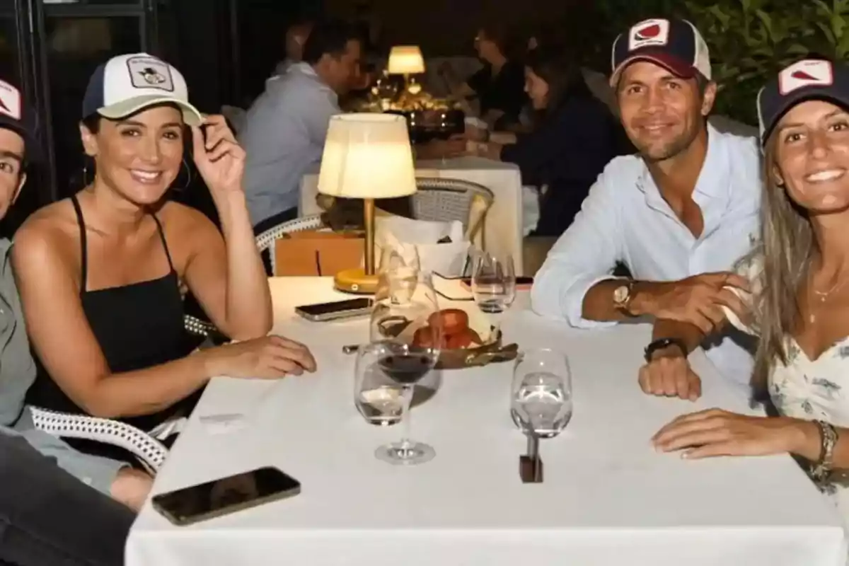 Un grupo de personas con gorras sentadas alrededor de una mesa en un restaurante, disfrutando de una cena con copas de vino y sonriendo a la cámara.