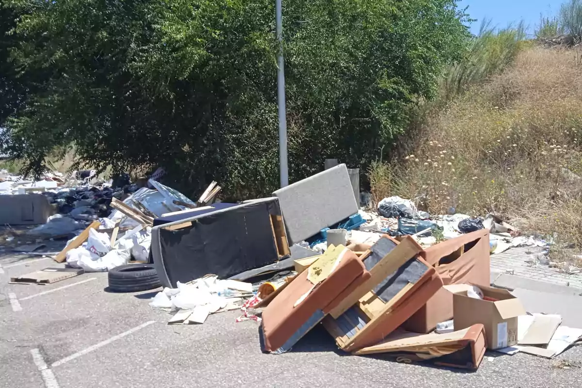 La imagen muestra una gran cantidad de basura y escombros, incluyendo muebles rotos, cajas de cartón, neumáticos y otros desechos, acumulados en una zona al aire libre cerca de un área con vegetación.