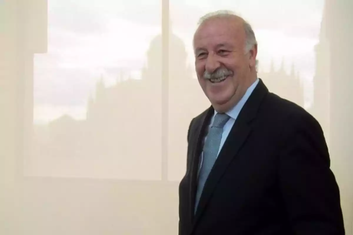 Un hombre mayor con bigote y traje oscuro sonríe frente a una ventana con una vista borrosa de un edificio al fondo.