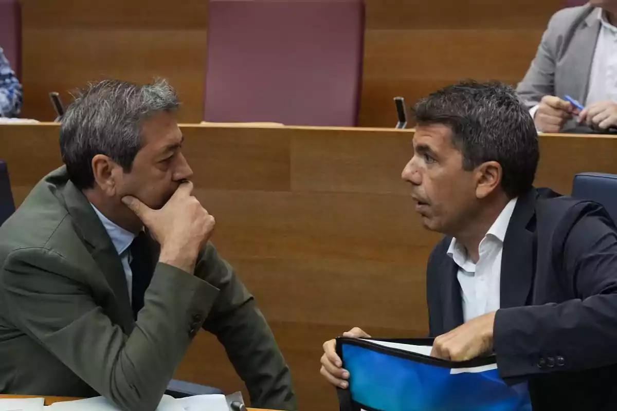 Dos hombres conversando en una sala de reuniones, uno de ellos sostiene una carpeta azul.