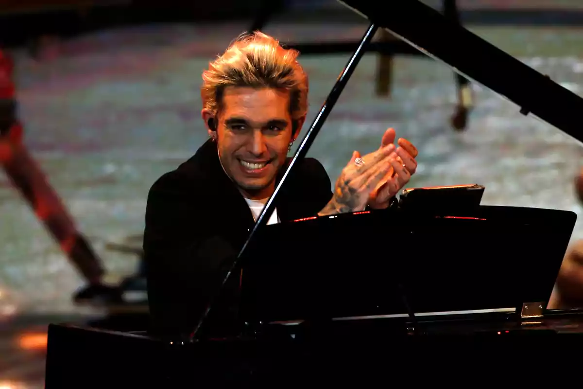 Un hombre con cabello rubio y sonrisa toca el piano mientras aplaude en un escenario iluminado.