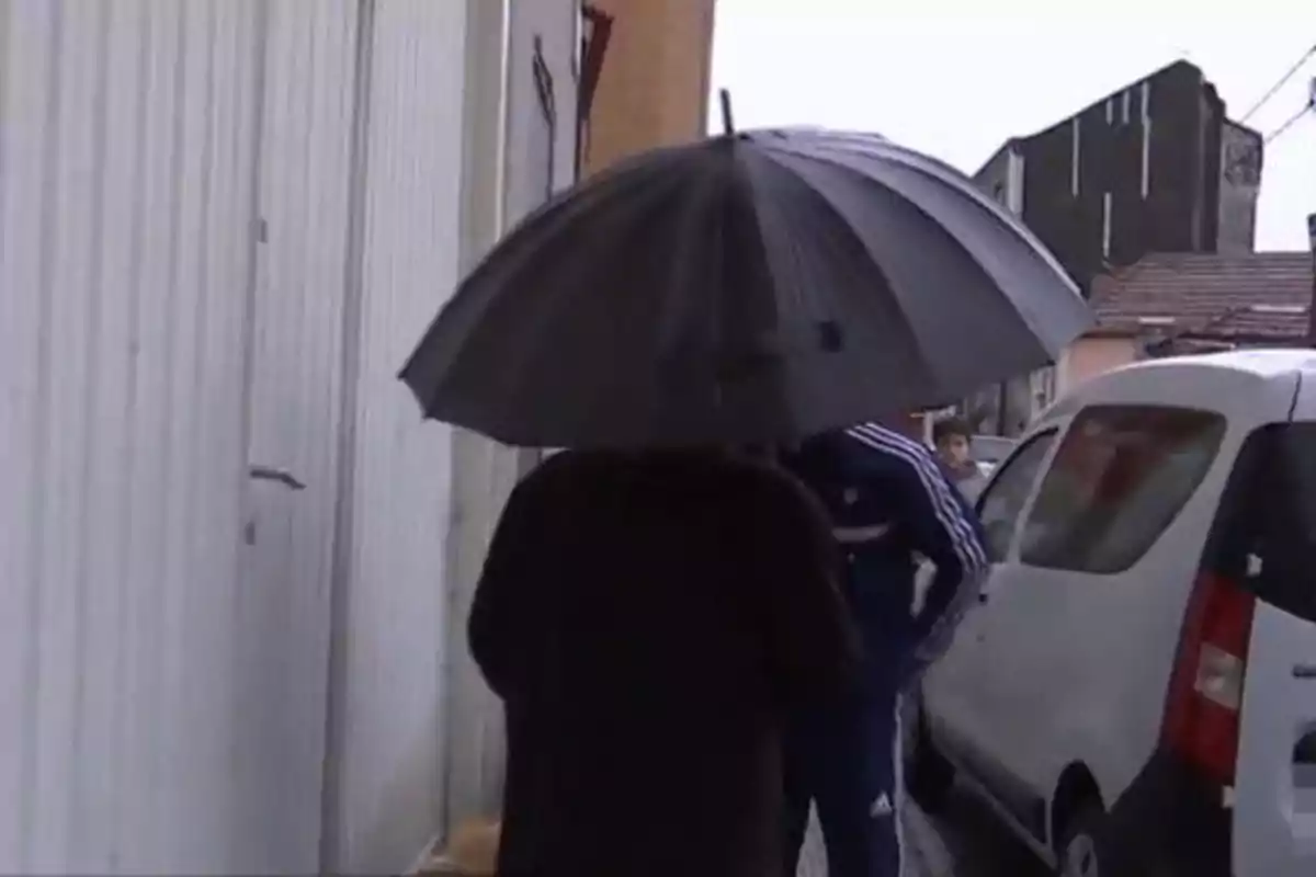 Persona con paraguas negro caminando por una calle junto a un edificio y un automóvil blanco.