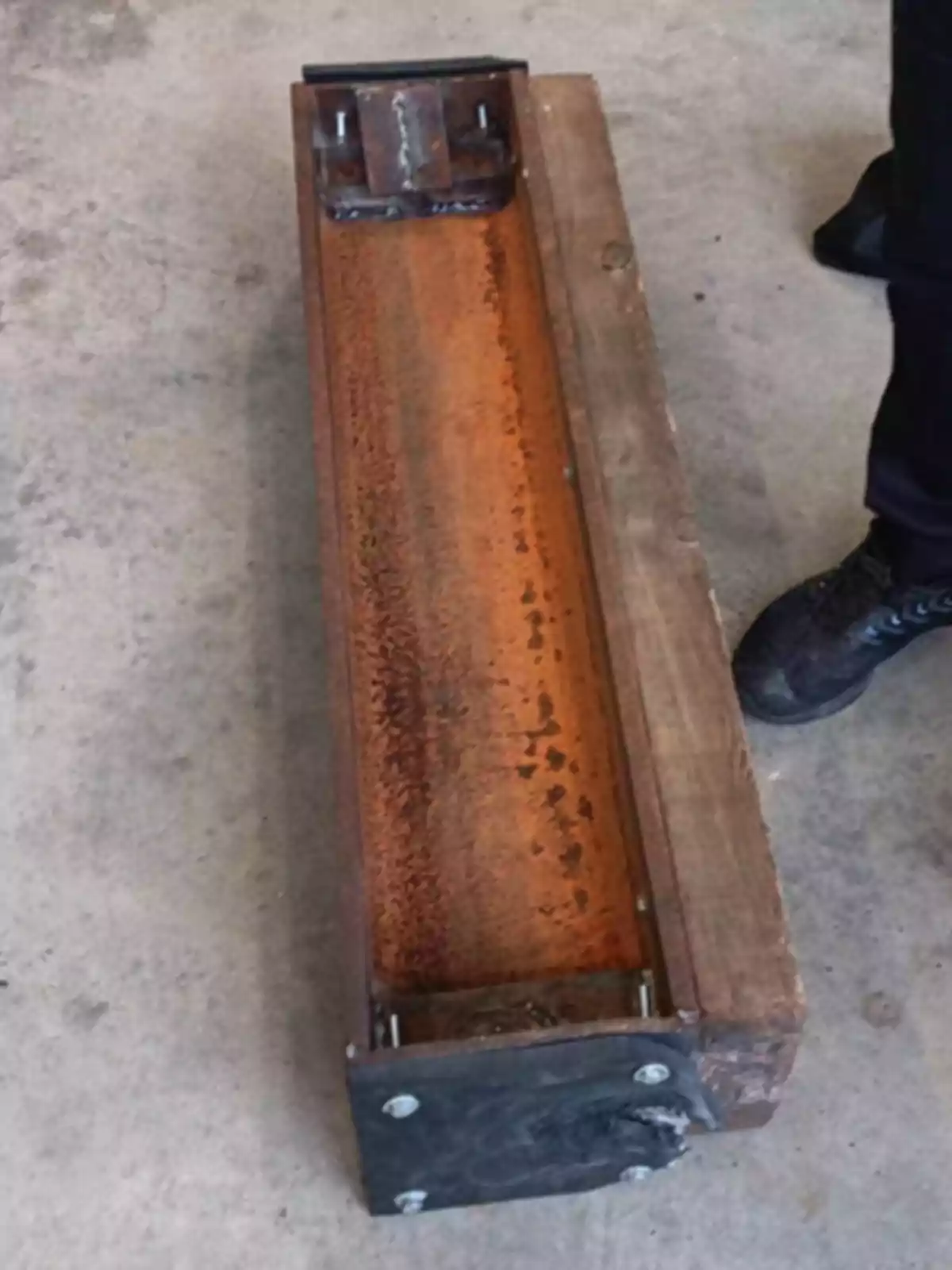 Un objeto rectangular de metal oxidado y madera sobre un suelo de concreto con una persona de pie al lado.