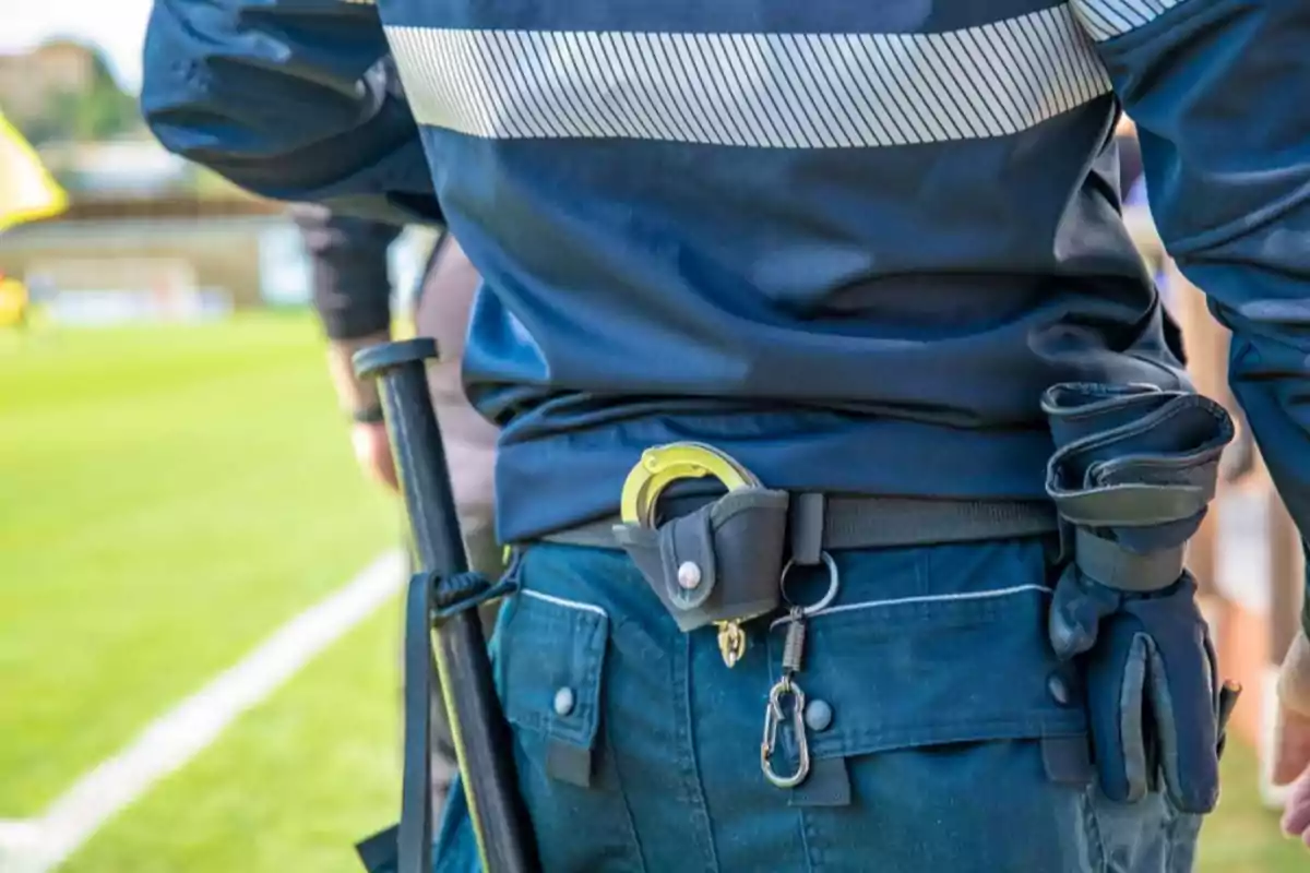 Persona con uniforme policial de espaldas en un campo de césped llevando esposas y un bastón en el cinturón.