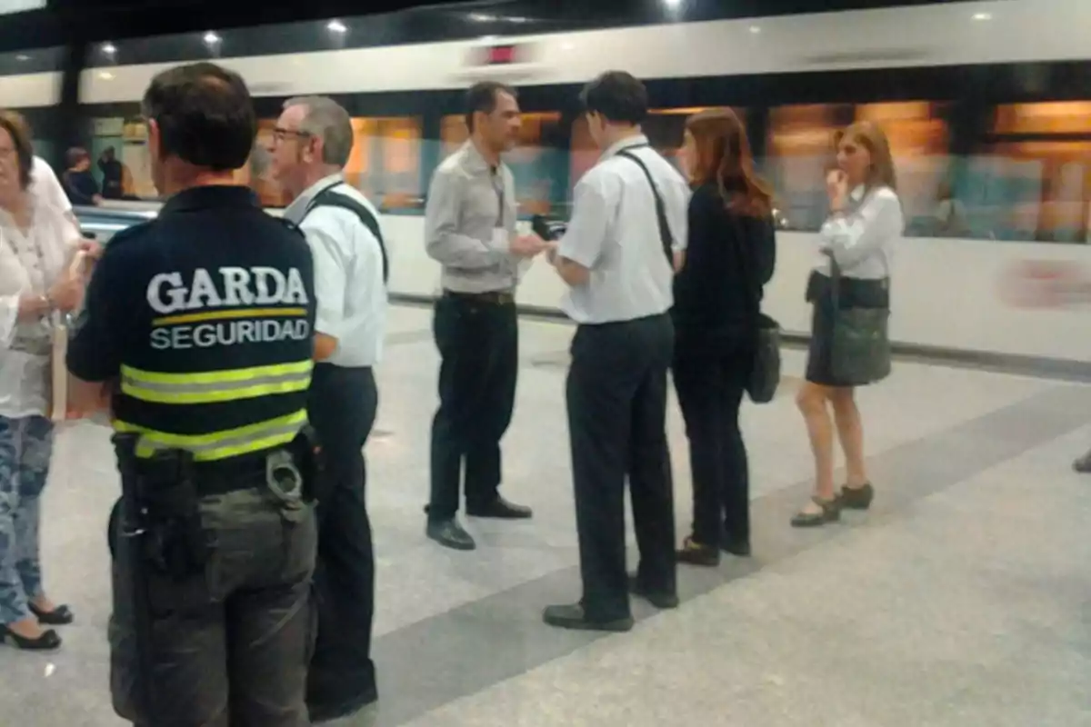 Un grupo de personas conversando en una estación de tren con un guardia de seguridad presente.