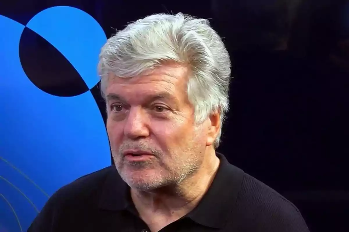 Hombre de cabello canoso y barba incipiente con camisa negra frente a un fondo azul y negro.