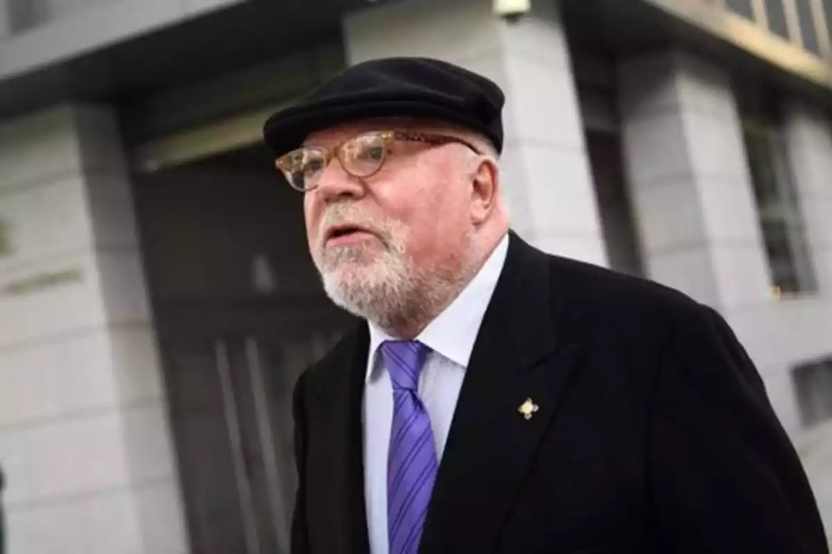 Hombre mayor con barba y gafas, vestido con traje oscuro, corbata morada y boina negra, caminando frente a un edificio moderno Hombre mayor con barba y gafas, vestido con traje oscuro, corbata morada y boina negra, caminando frente a un edificio moderno