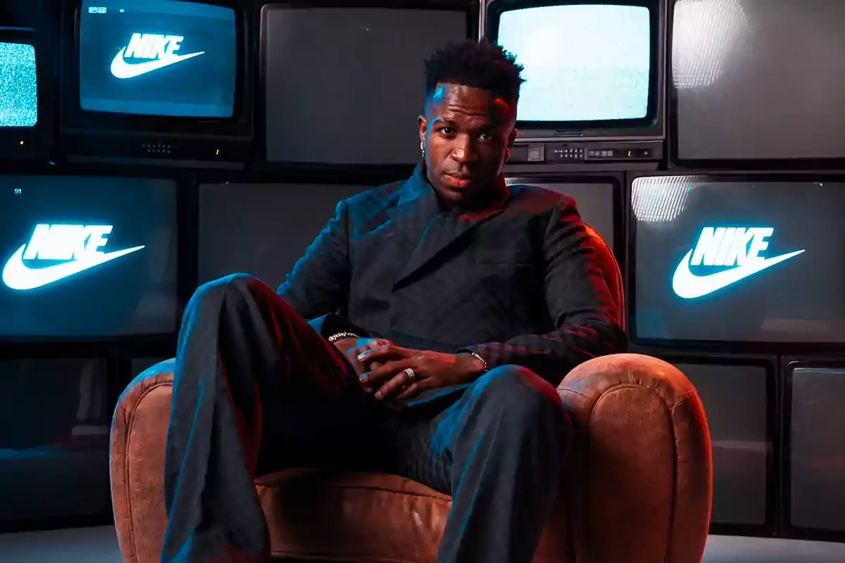 Un hombre sentado en un sillón frente a una pared de televisores antiguos que muestran el logotipo de Nike. Un hombre sentado en un sillón frente a una pared de televisores antiguos que muestran el logotipo de Nike.