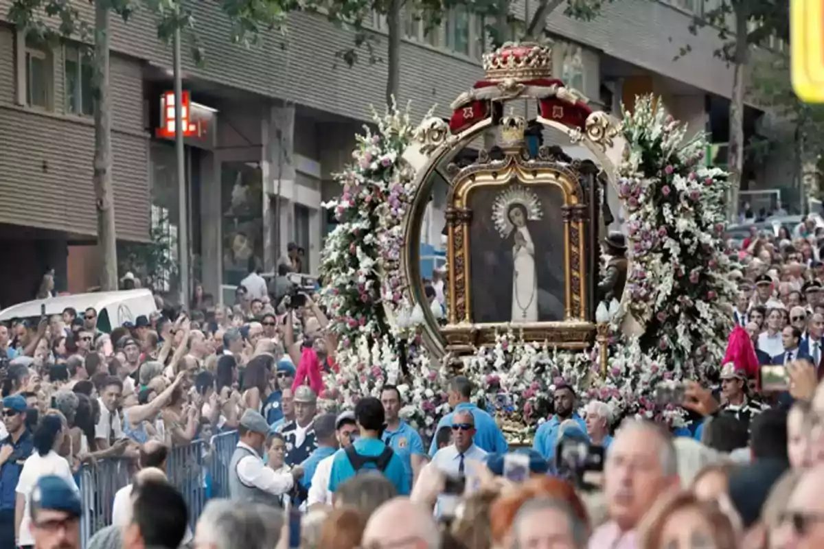 Una multitud de personas participa en una procesión religiosa, llevando una imagen sagrada adornada con flores y rodeada de devotos.