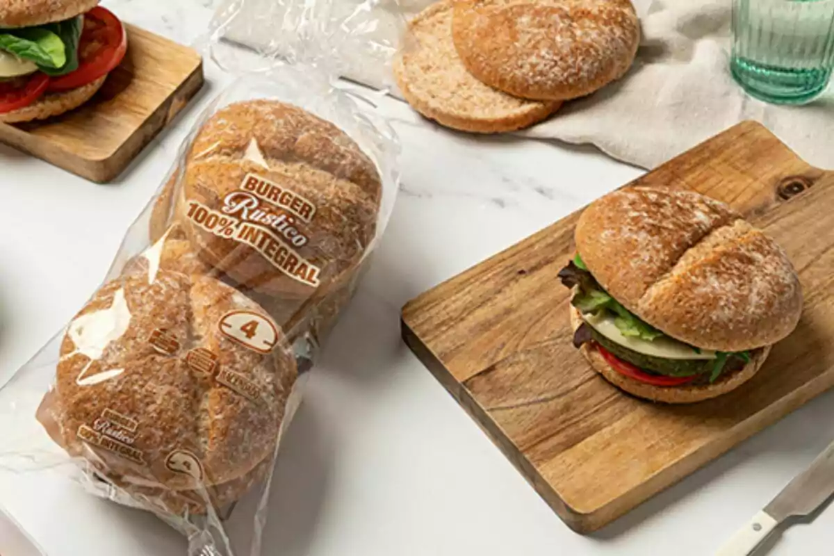 Un paquete de pan rústico integral junto a una hamburguesa con ingredientes frescos sobre una tabla de madera. Un paquete de pan rústico integral junto a una hamburguesa con ingredientes frescos sobre una tabla de madera.