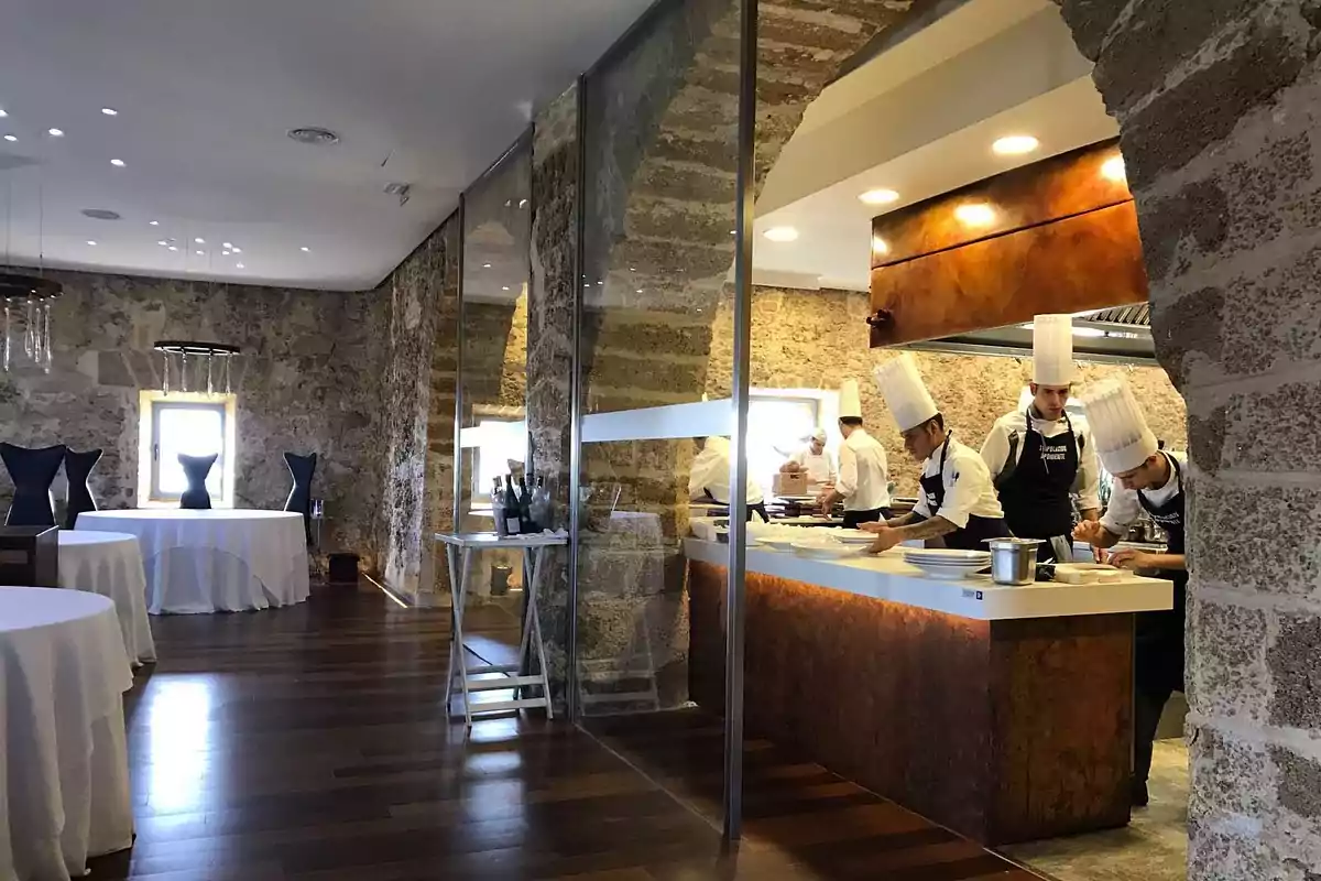 Cocina de un restaurante con chefs trabajando, mesas redondas con manteles blancos y sillas negras en un ambiente de paredes de piedra y suelo de madera.