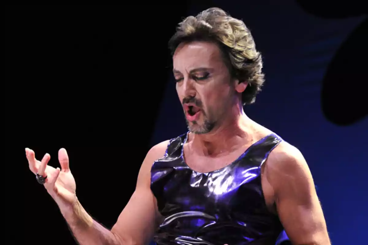 Hombre con barba corta y maquillaje escénico cantando apasionadamente en un escenario con fondo oscuro