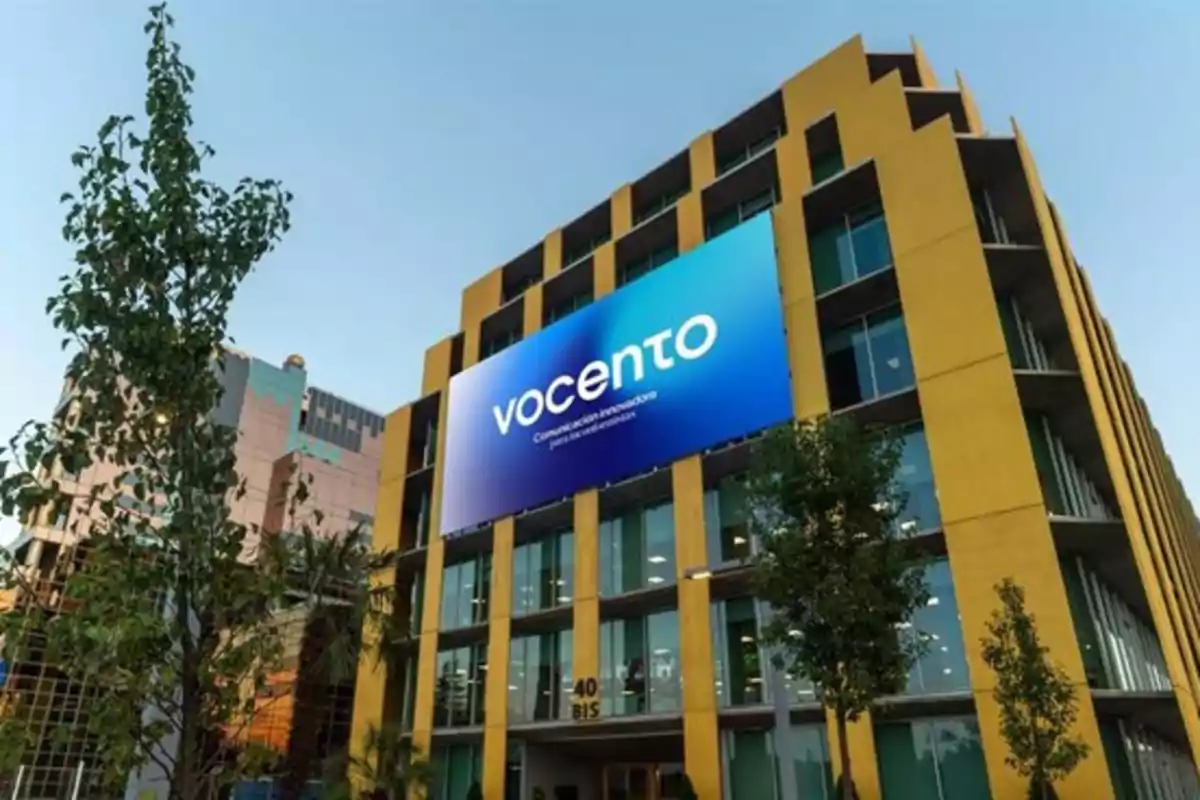 Edificio moderno de color amarillo con un gran cartel azul que muestra el logo de vocento rodeado de árboles y cielo despejado Edificio moderno de color amarillo con un gran cartel azul que muestra el logo de vocento rodeado de árboles y cielo despejado