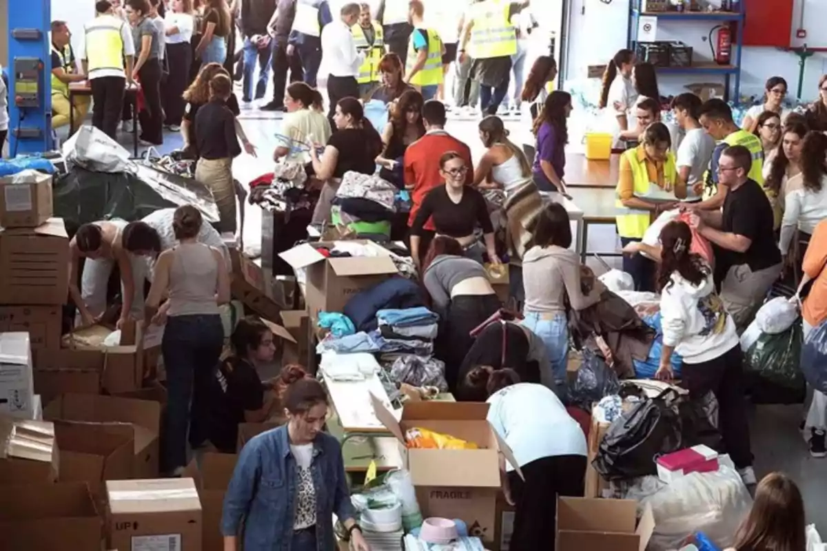 Un grupo de personas organiza y clasifica ropa y otros artículos en un centro de acopio, rodeados de cajas y bolsas.