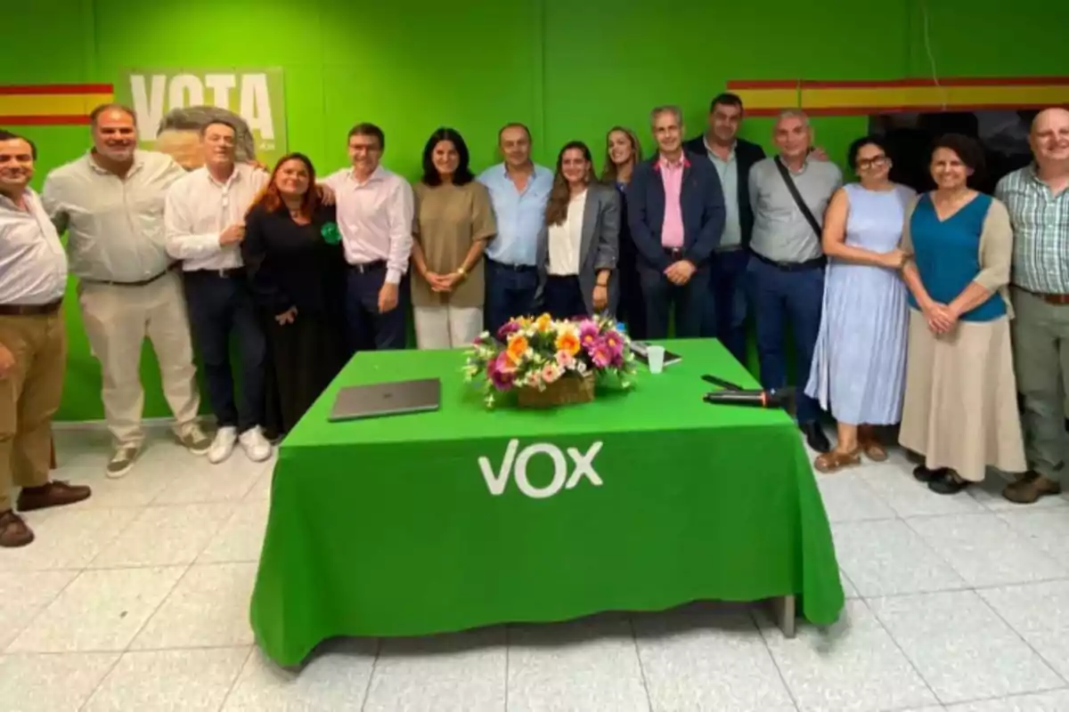 Un grupo de personas posando en una sala con paredes verdes y una mesa con un mantel verde que tiene el logo de VOX, adornada con un arreglo floral.