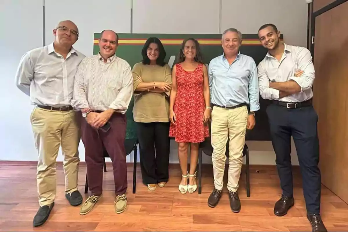 Un grupo de seis personas posando juntas en una sala con piso de madera y una pizarra al fondo.