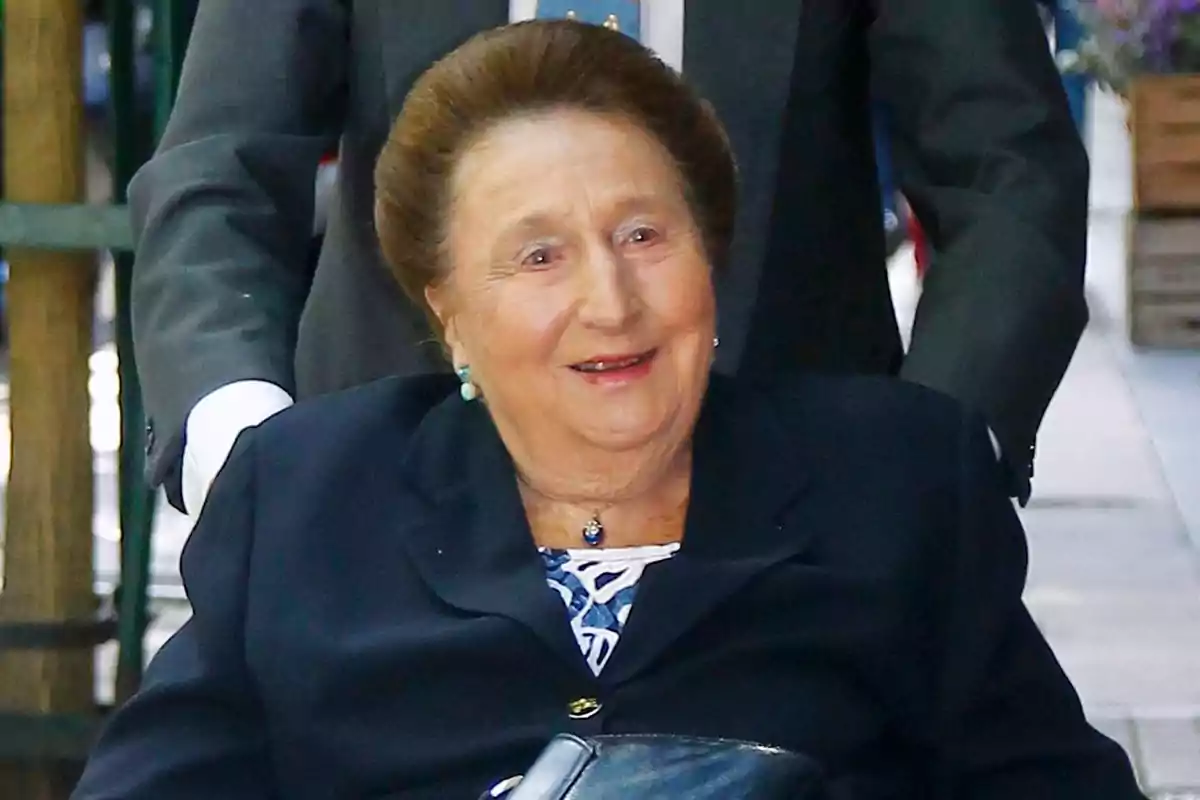 Mujer mayor sonriente vestida con saco oscuro y blusa estampada siendo empujada en una silla de ruedas por una persona con traje en un entorno urbano Mujer mayor sonriente vestida con saco oscuro y blusa estampada siendo empujada en una silla de ruedas por una persona con traje en un entorno urbano