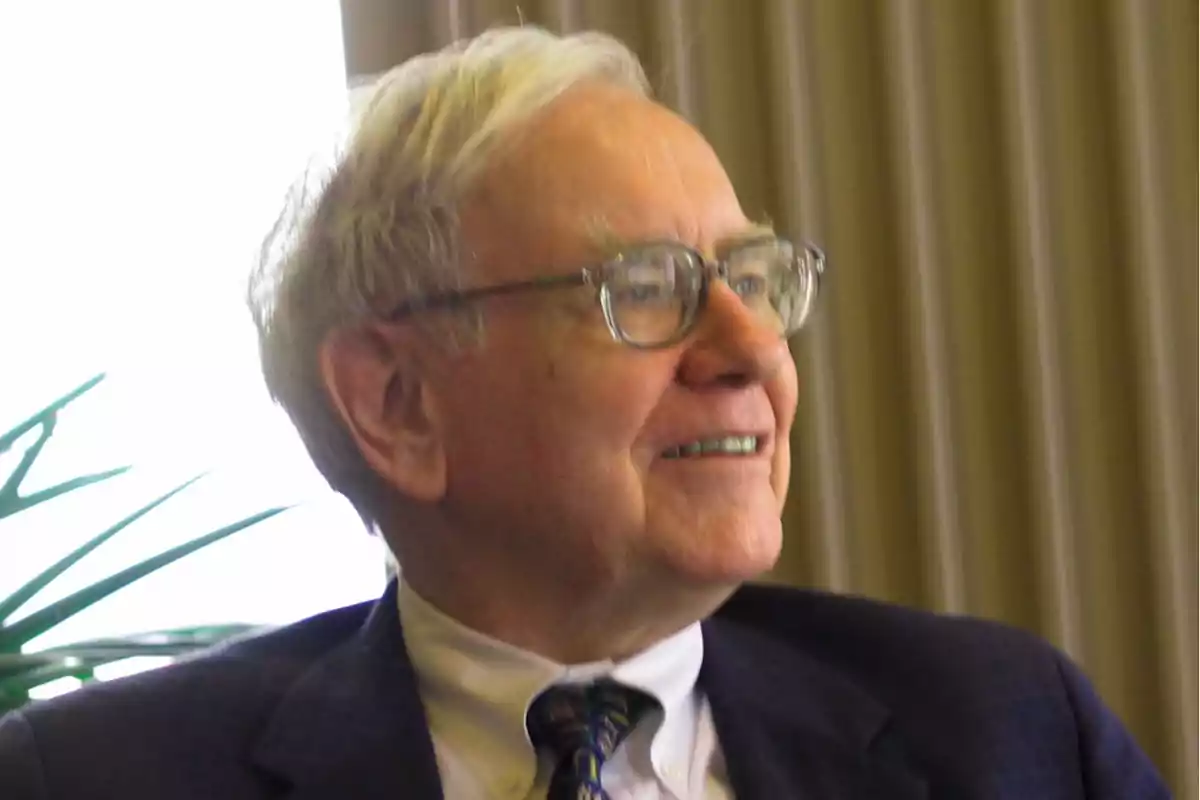 An older man with glasses and gray hair, smiling and wearing a dark suit with a tie, sitting in an indoor setting. An older man with glasses and gray hair, smiling and wearing a dark suit with a tie, sitting in an indoor setting.