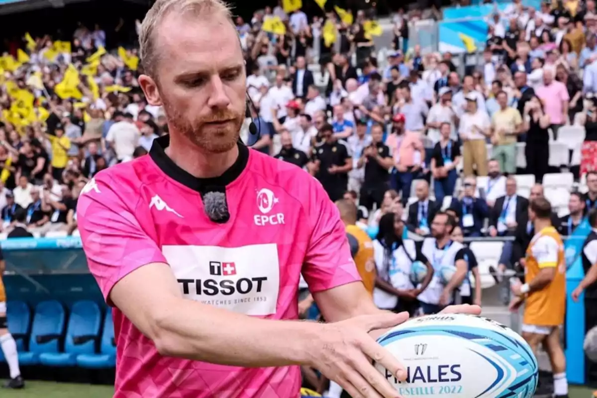 Un árbitro de rugby con uniforme rosa sostiene un balón mientras se prepara para un partido, con una multitud de espectadores en el fondo.