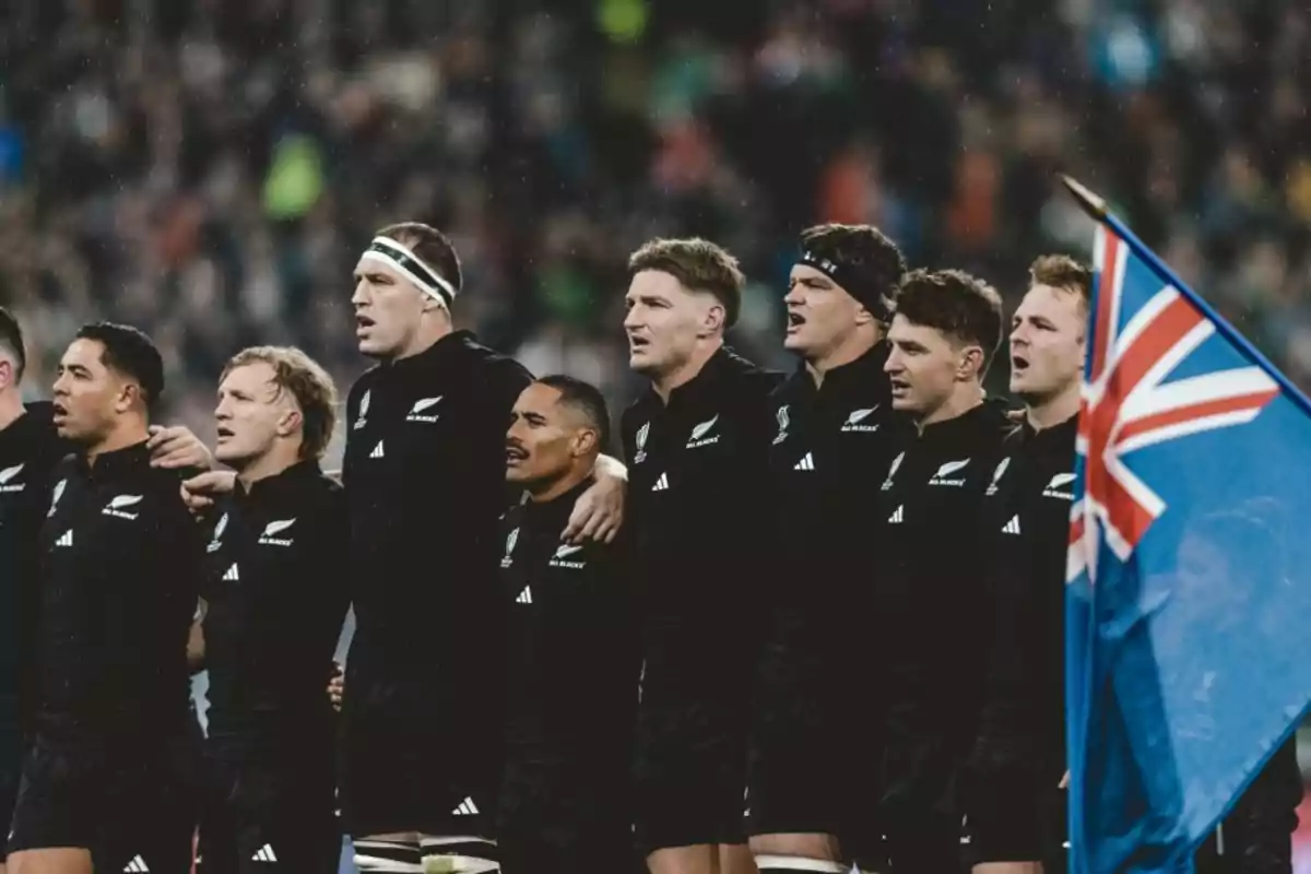 Jugadores de rugby en uniforme negro alineados y abrazados mientras cantan, con una bandera azul y roja en primer plano.