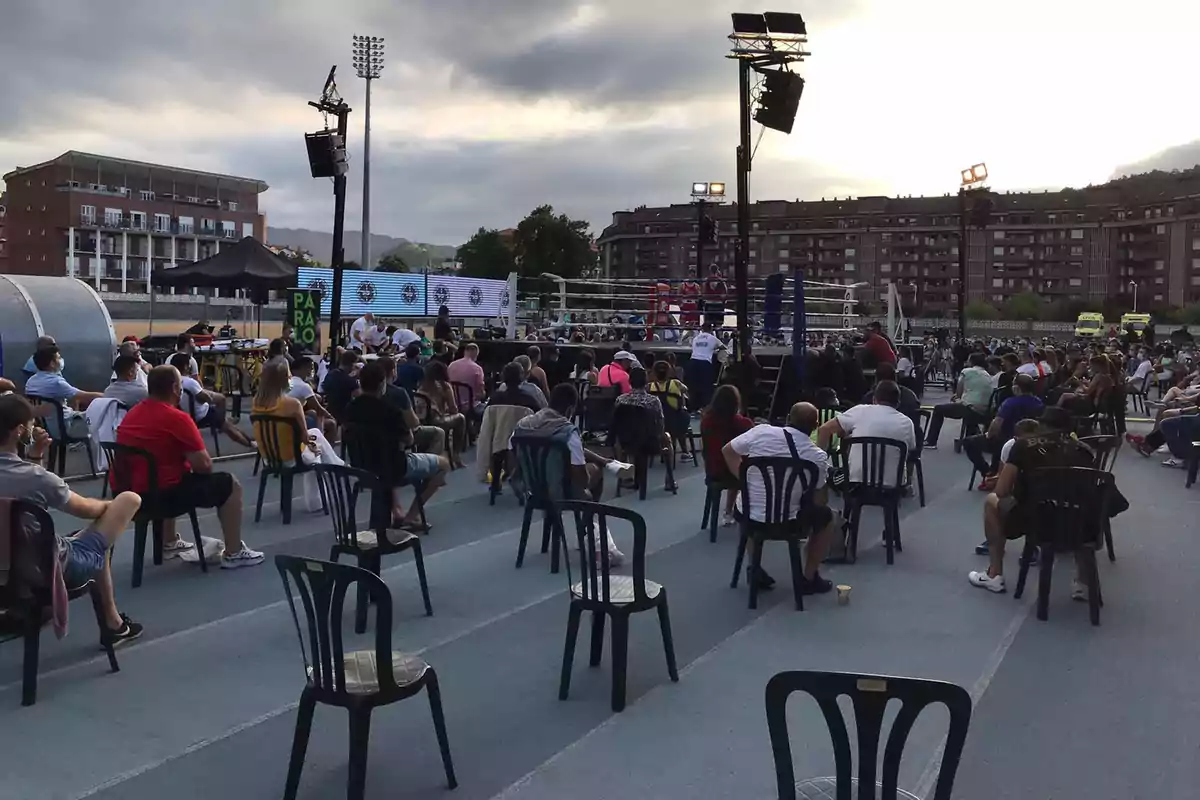 Una multitud de personas sentadas en sillas al aire libre observando un evento de boxeo en un ring, con edificios y luces de estadio en el fondo.