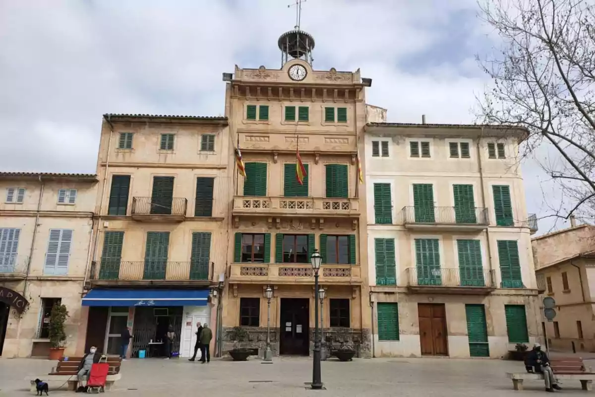 Fachada de un edificio histórico con ventanas de contraventanas verdes, un reloj en la parte superior y varias banderas en el balcón, situado en una plaza con bancos y algunas personas Fachada de un edificio histórico con ventanas de contraventanas verdes, un reloj en la parte superior y varias banderas en el balcón, situado en una plaza con bancos y algunas personas