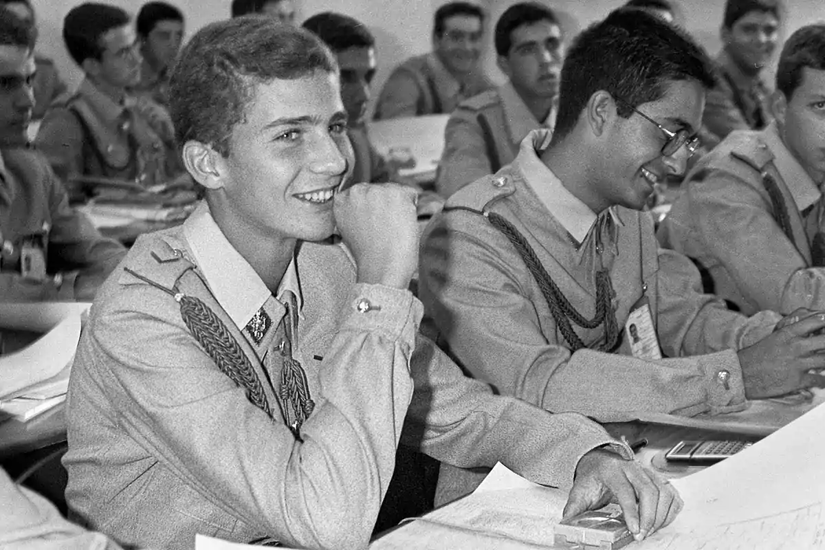 Un grupo de jóvenes cadetes en uniforme militar asisten a una clase, con uno de ellos sonriendo y apoyando su brazo en la mesa.