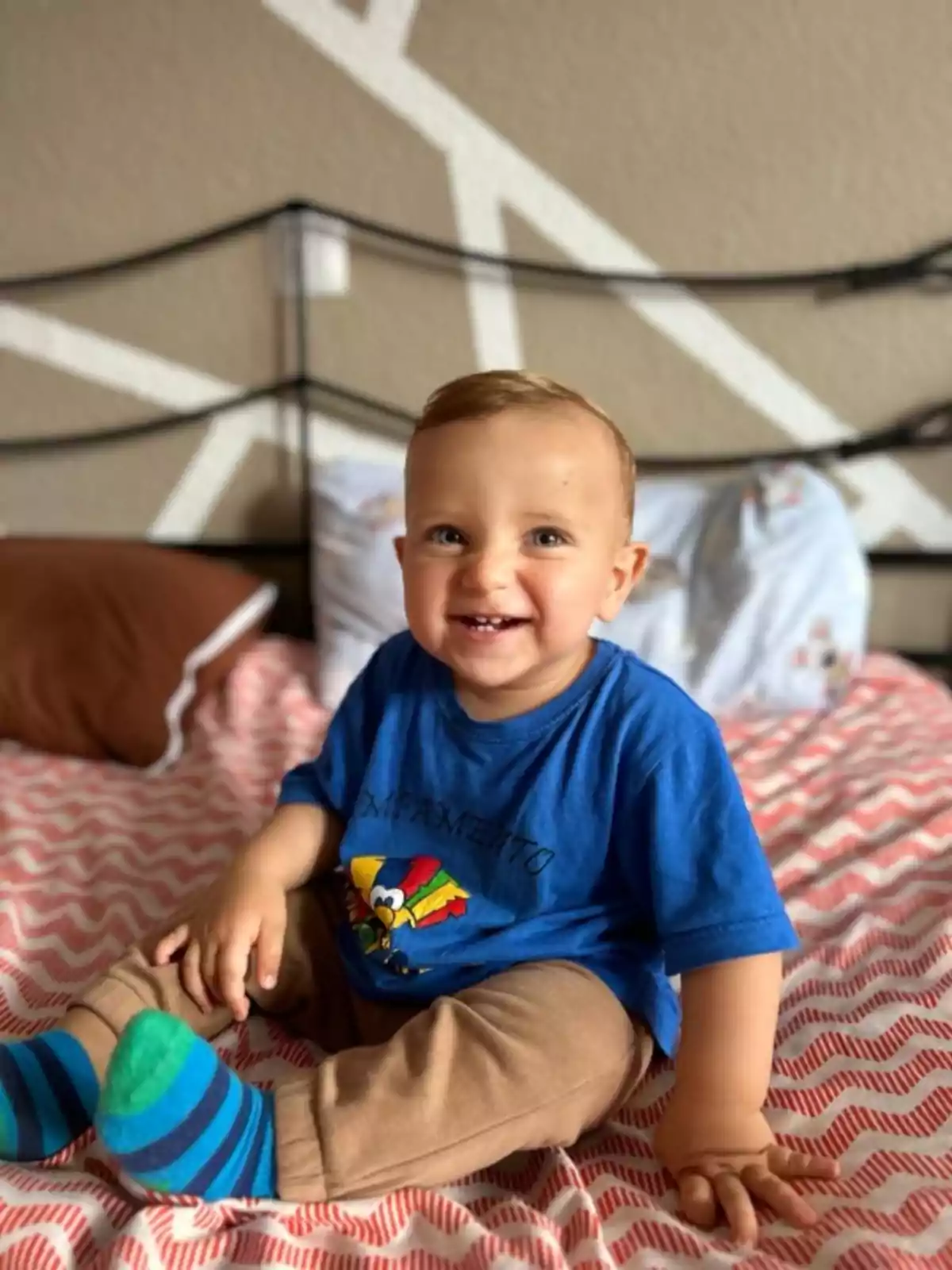 Un niño pequeño sonriente sentado en una cama con una camiseta azul y calcetines a rayas.