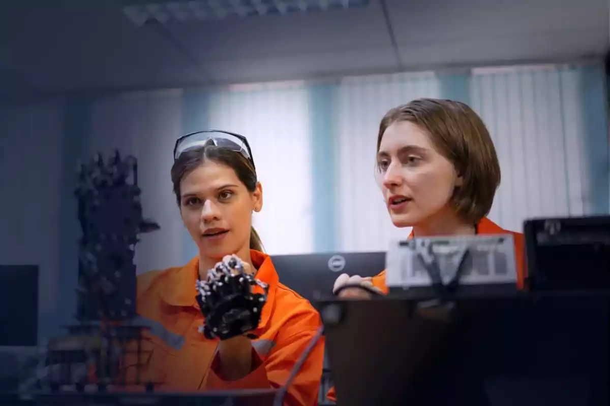 Dos mujeres en un laboratorio observando y discutiendo sobre un dispositivo mecánico mientras usan uniformes naranjas.
