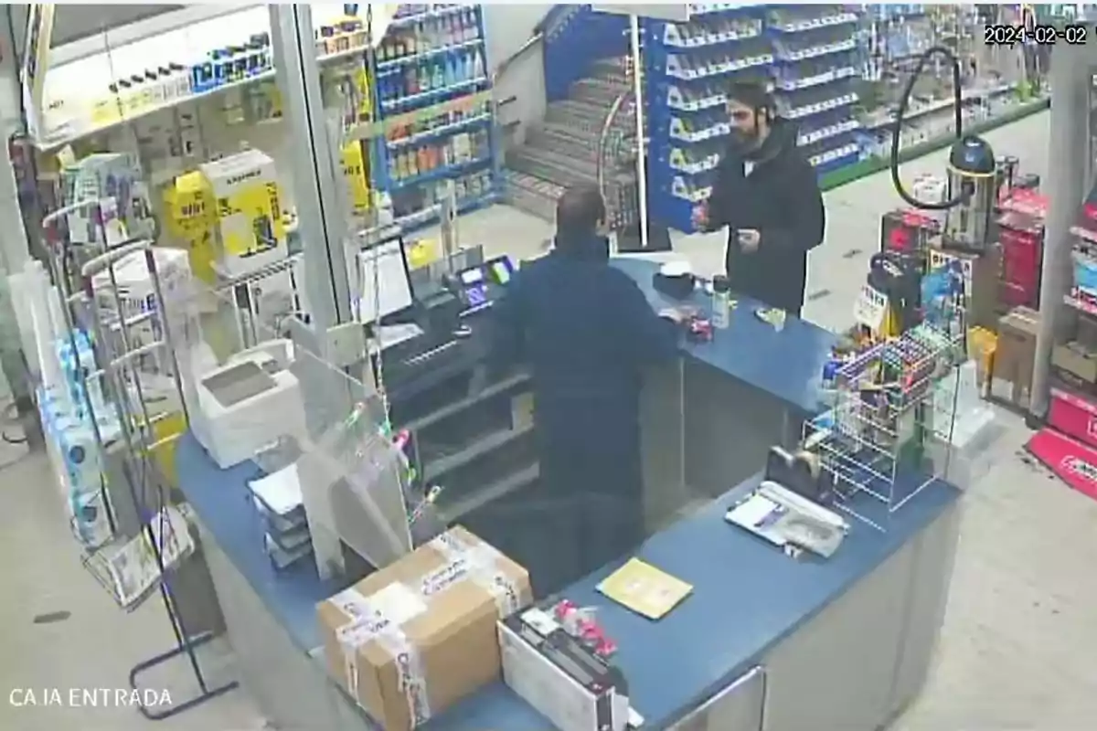 Una persona con barba está hablando con un empleado detrás de un mostrador en una tienda, rodeados de estantes con productos y cajas en el mostrador.