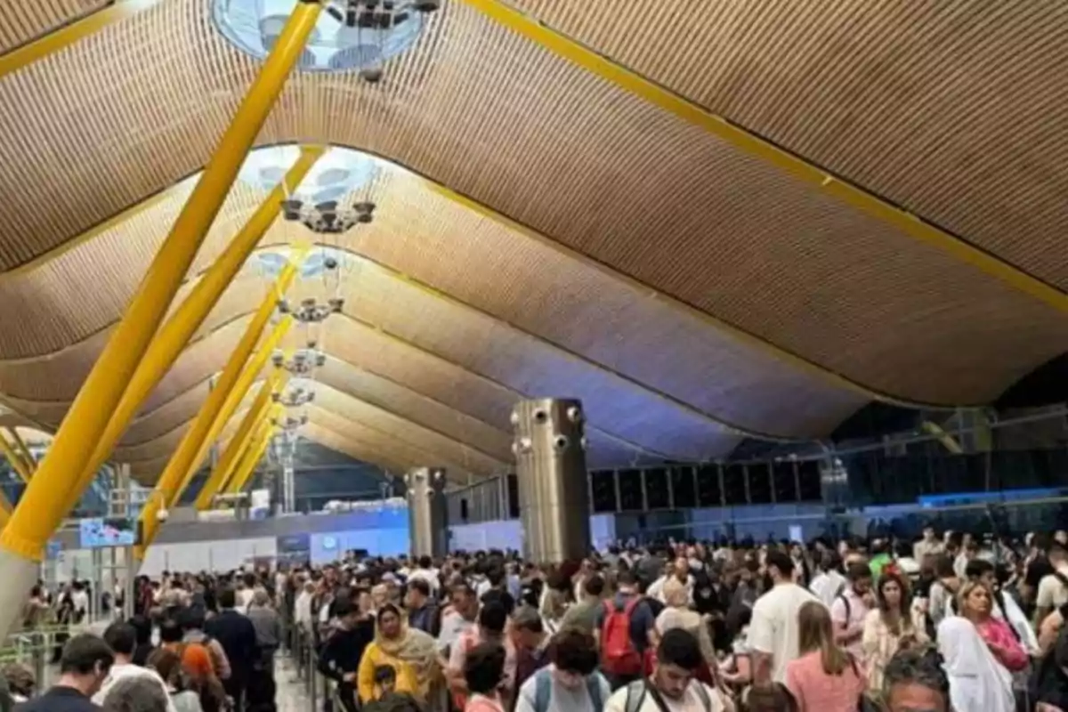 Una multitud de personas en el interior de un aeropuerto con un techo de diseño moderno y columnas amarillas.