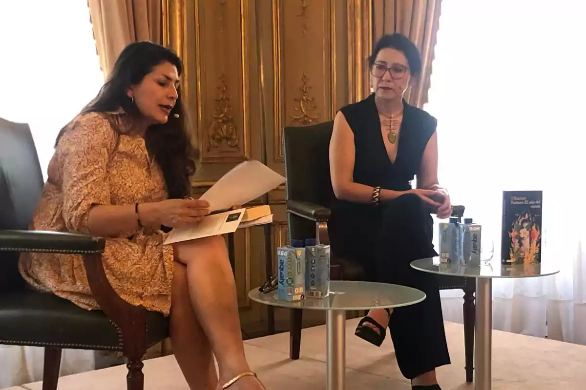 Dos mujeres sentadas en sillas durante una presentación, una de ellas leyendo un documento mientras la otra escucha atentamente, con una mesa de vidrio entre ellas que sostiene cajas de agua y un libro.