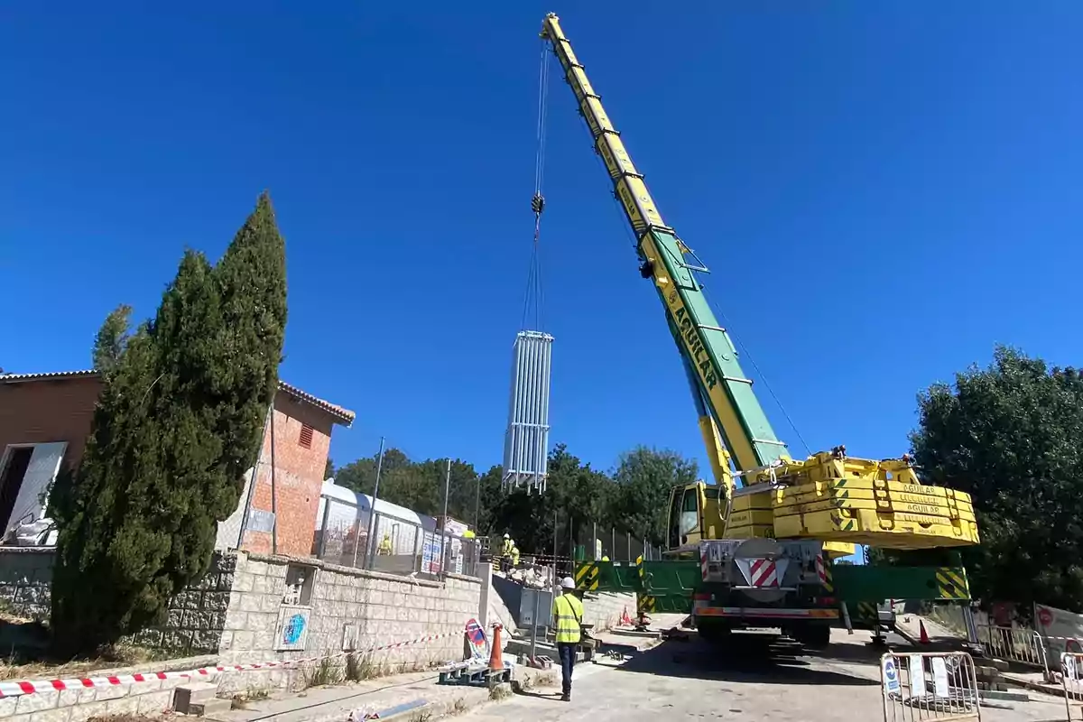 Una grúa levantando una estructura metálica en un sitio de construcción con trabajadores y árboles alrededor.