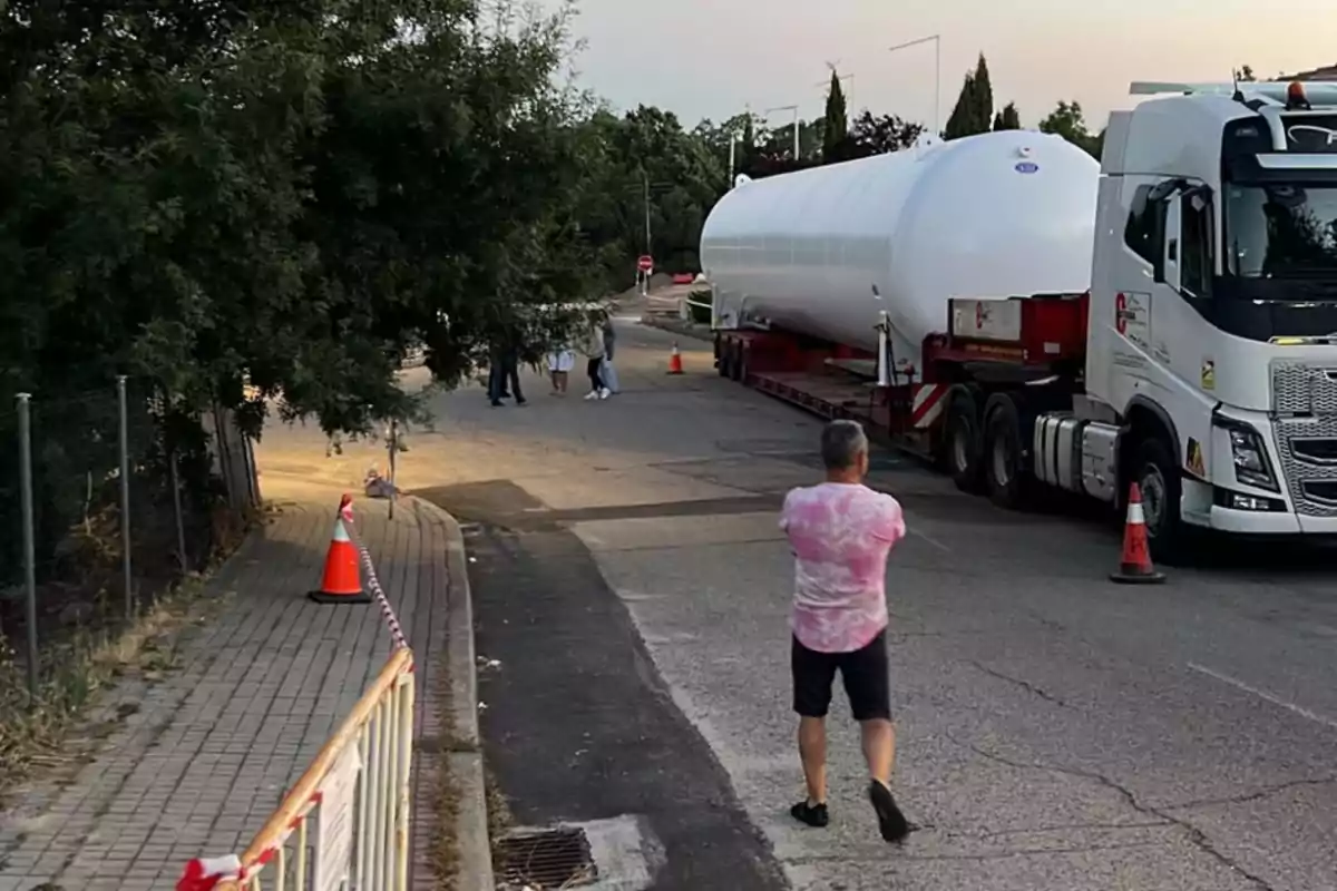 Un hombre con camiseta rosa observa un camión que transporta un gran tanque blanco en una calle.