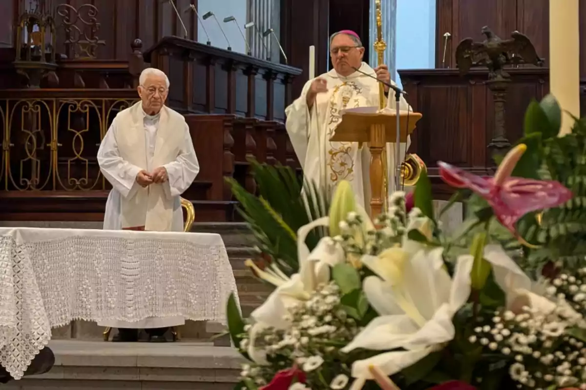 Dos sacerdotes oficiando una misa en una iglesia, con un altar decorado con flores en primer plano.